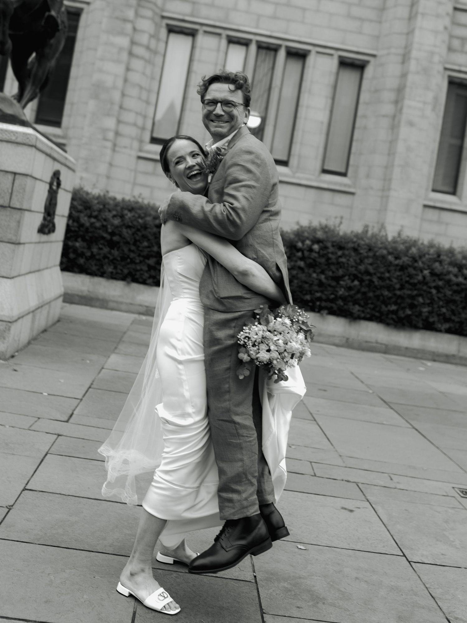 Black and white photo of a joyful couple embracing outdoors on their wedding day, bride in white dress holding bouquet.