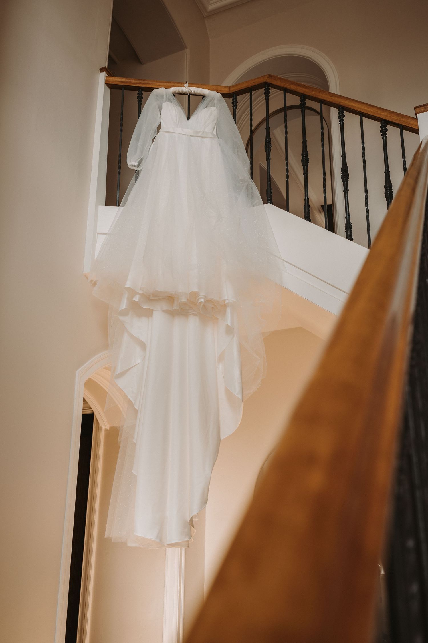 A white long-sleeved wedding dress hanging on a staircase banister with delicate tulle veil flowing down the stairs at Kinclune House.