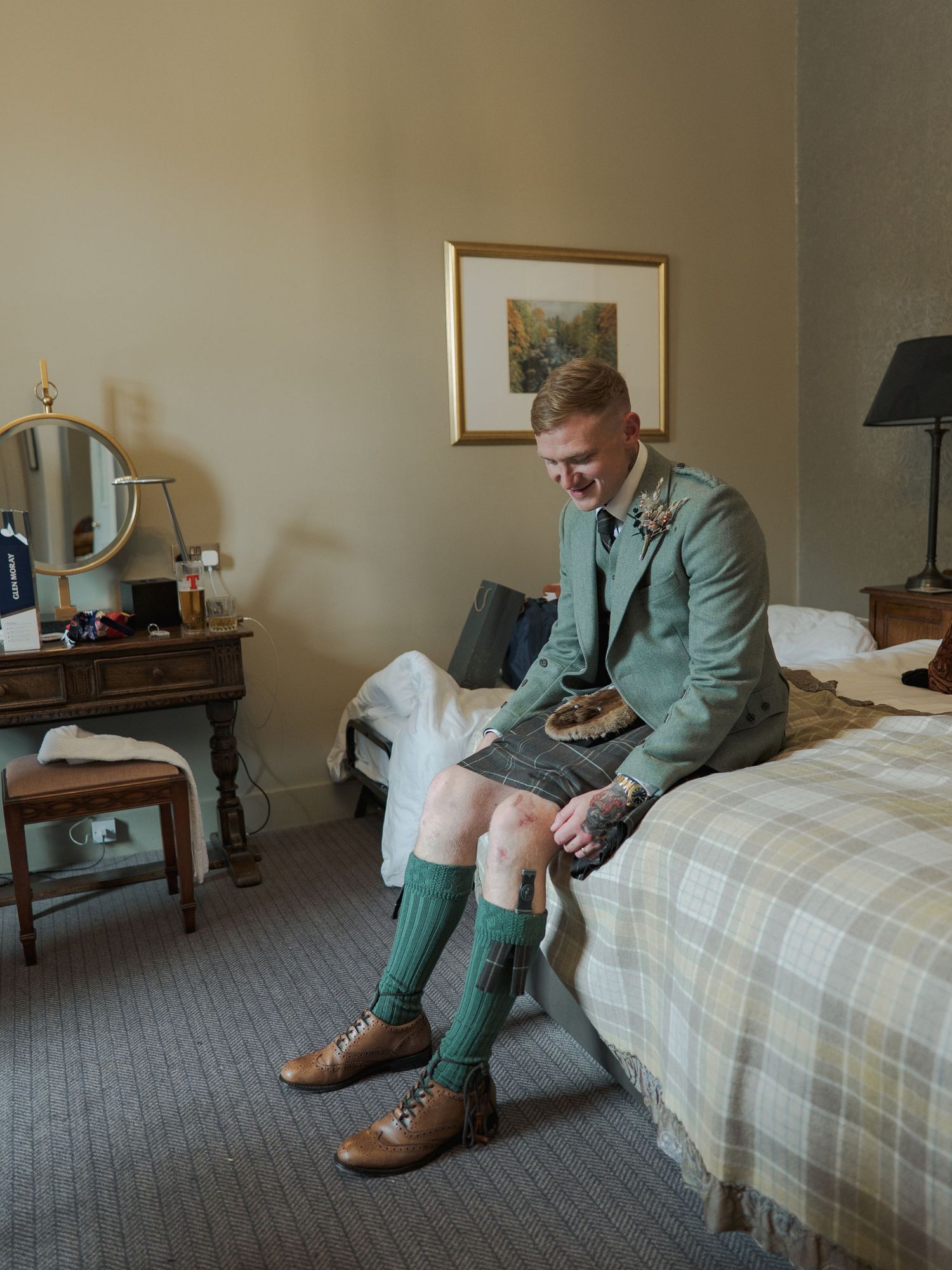 A person sits on a hotel bed wearing sage green attire and brown leather boots.