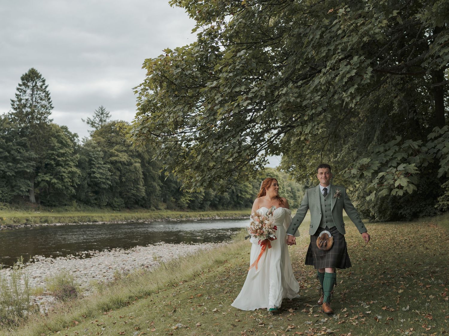 Wedding celebration group walks along scenic tree-lined path with overcast sky in background at Banchory Lodge.