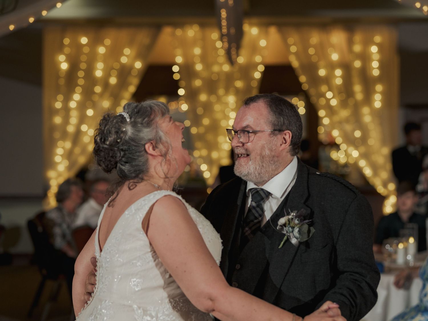 Elderly couple sharing a romantic first dance at their wedding reception with warm fairy lights glowing in the background.