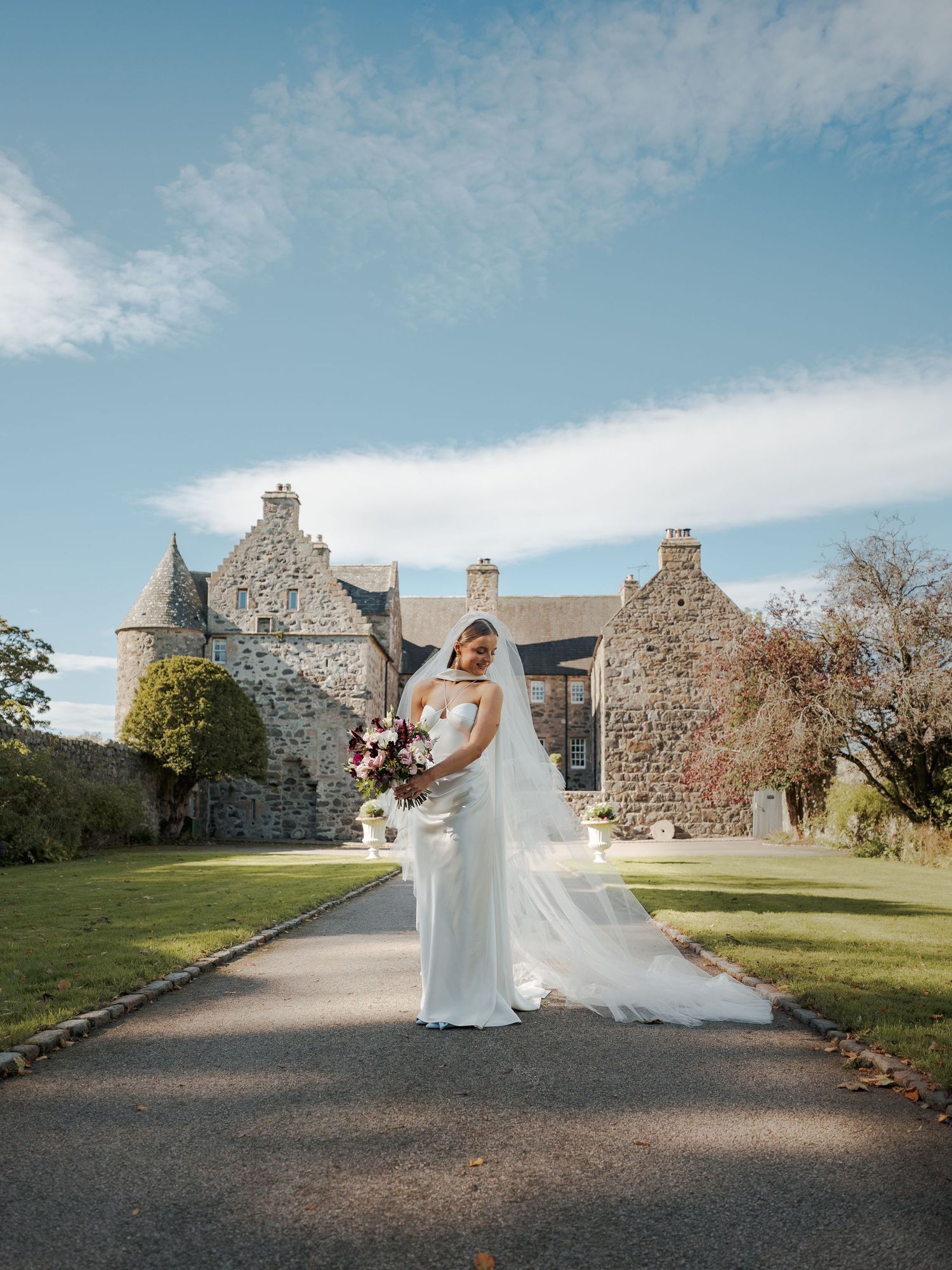 Bride in white gown and veil holds bouquet on path before Scottish stone castle under blue sky.
