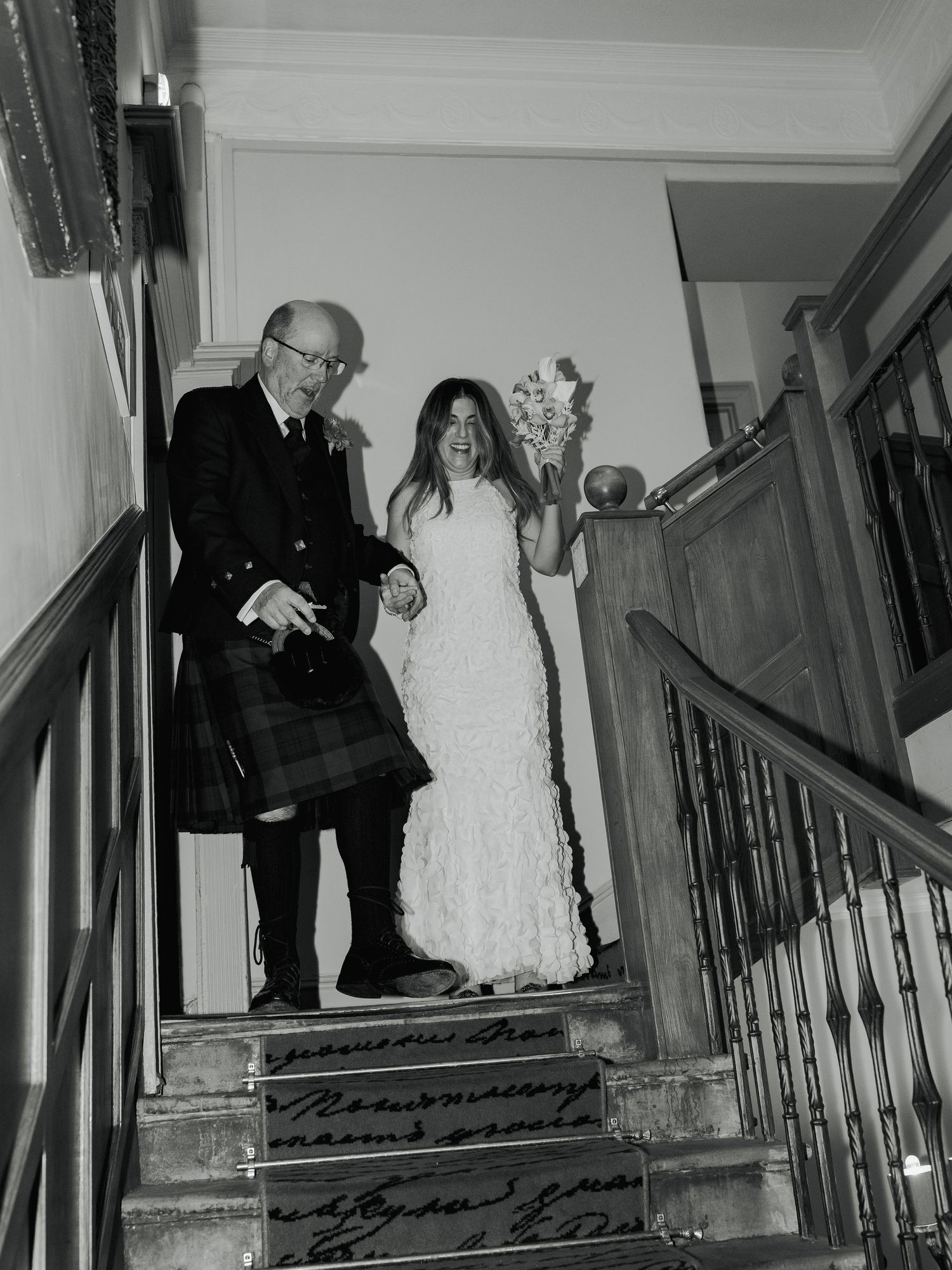 Bride in white dress holding bouquet walks down staircase with older man in Scottish tartan kilt.