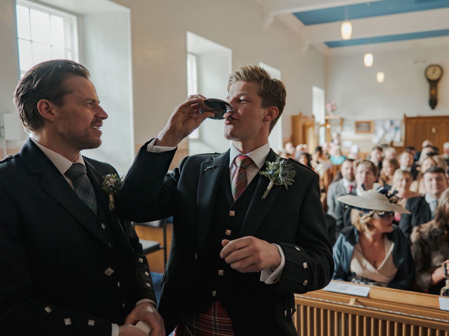 Two grooms share a toast at their wedding ceremony in a rustic venue, wearing kilts and boutonnieres.