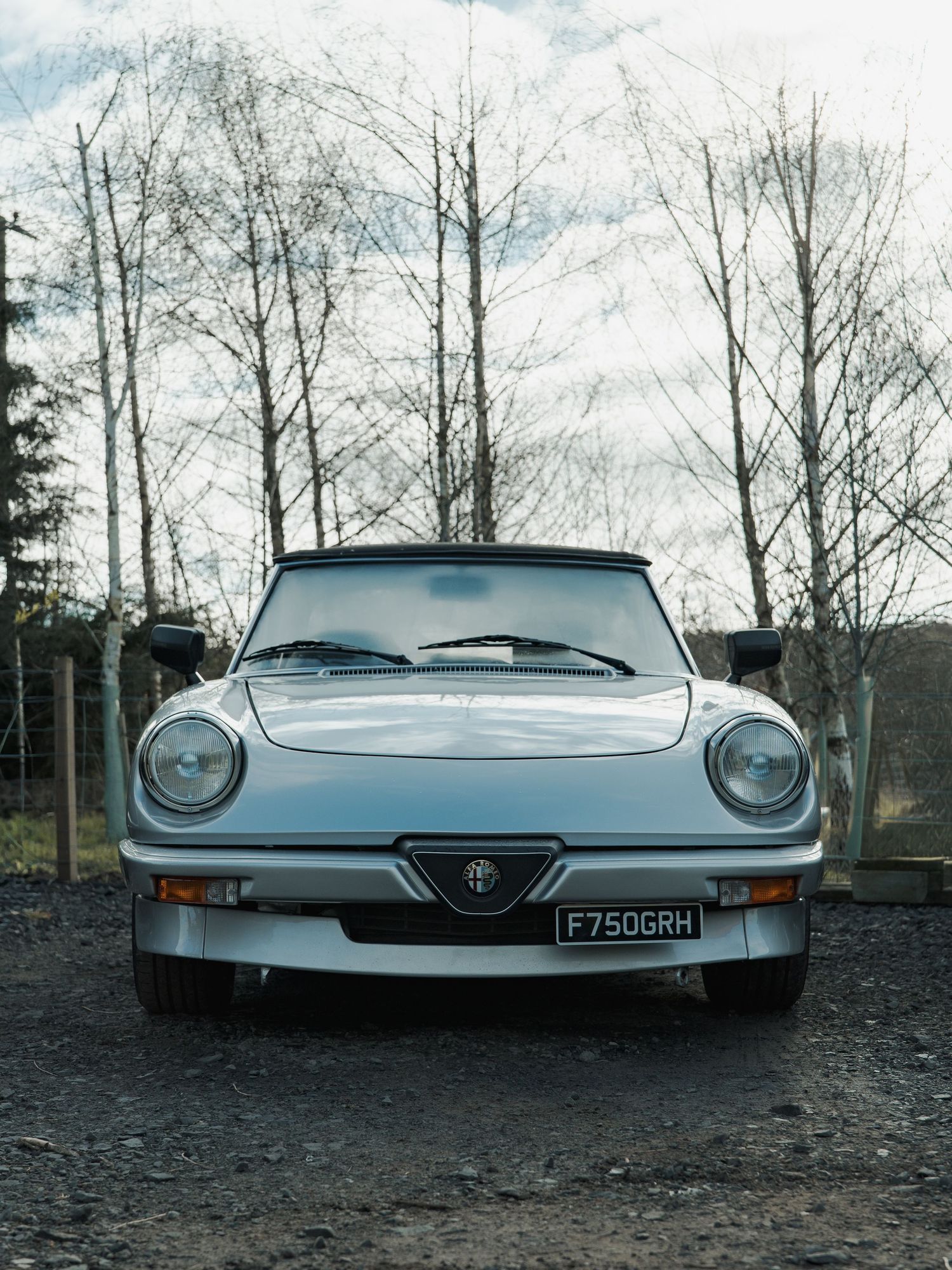 A classic silver Alfa Romeo Spider convertible parked on gravel, front view, UK registration plate F750GRH, bare trees background.