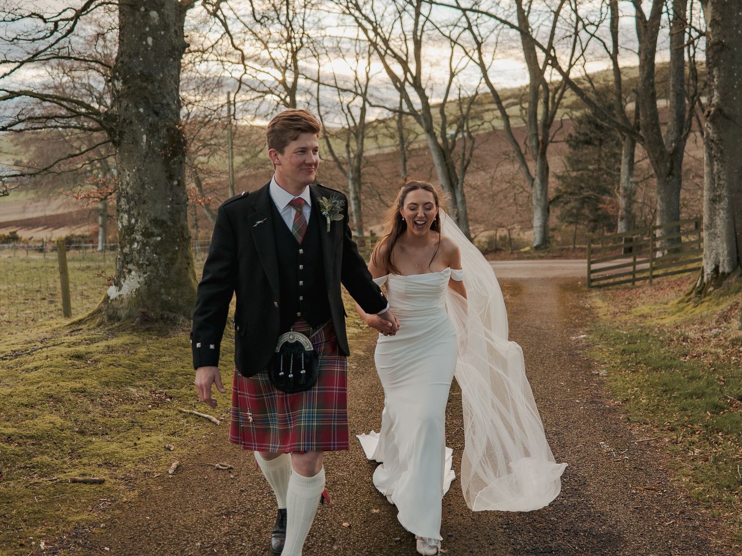 A groom in a Scottish kilt and bride in a white gown walk hand-in-hand along a tree-lined path at Kinclune House