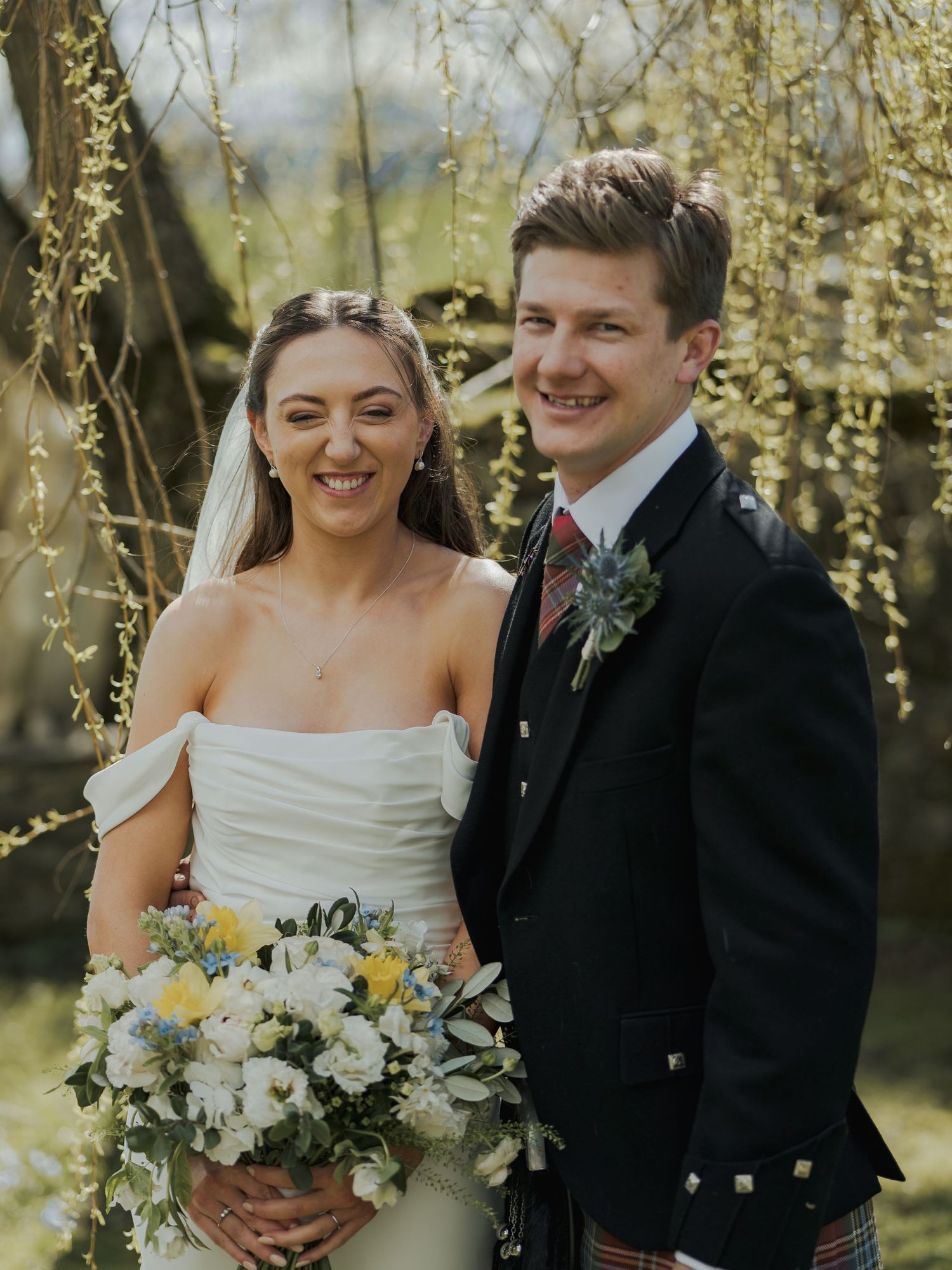Smiling bride and groom in Scottish kilt pose together outdoors under a weeping willow tree on their wedding day.