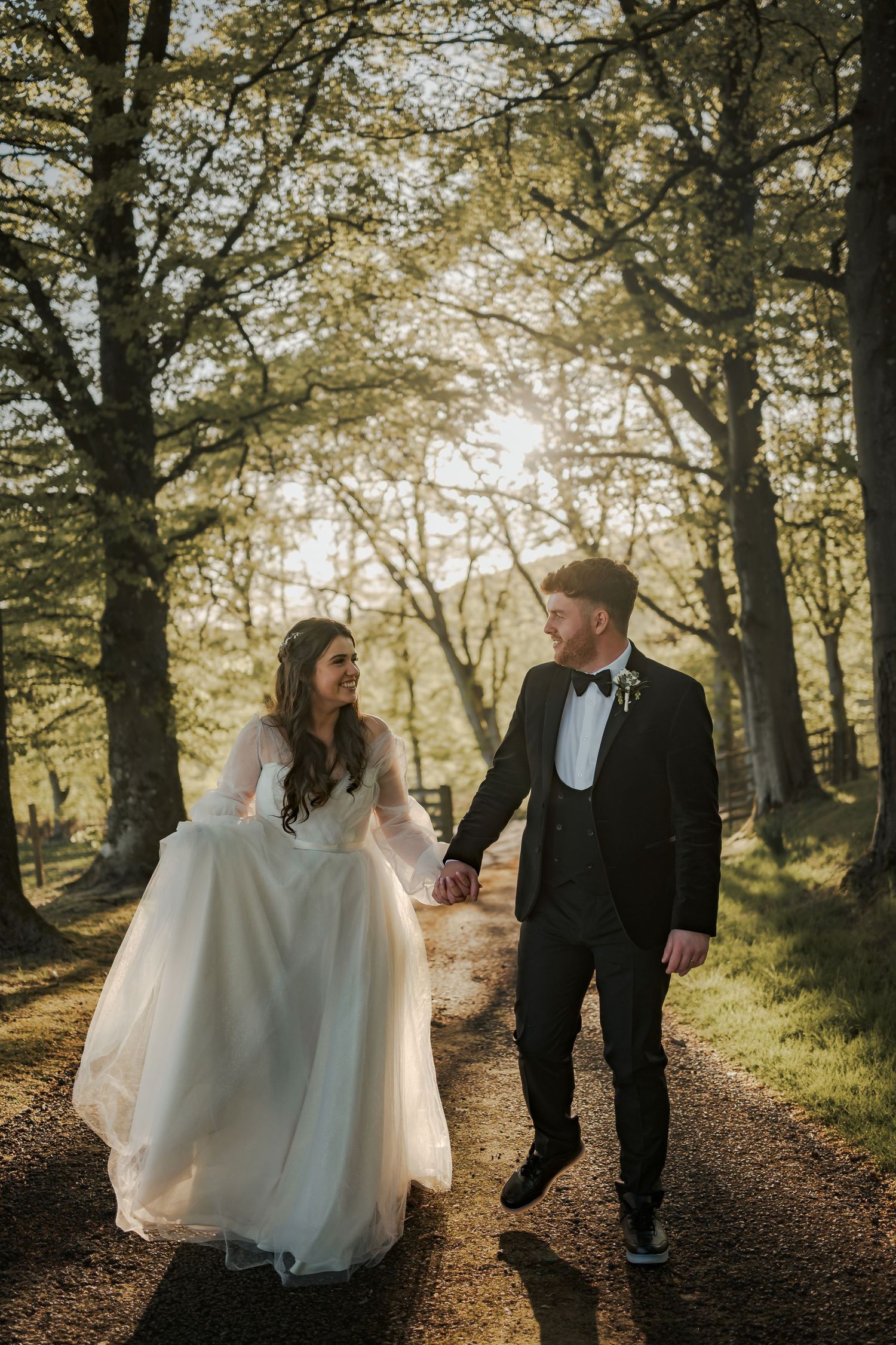 Bride and groom walk hand in hand along a sunlit woodland path, smiling at each other on their wedding day.
