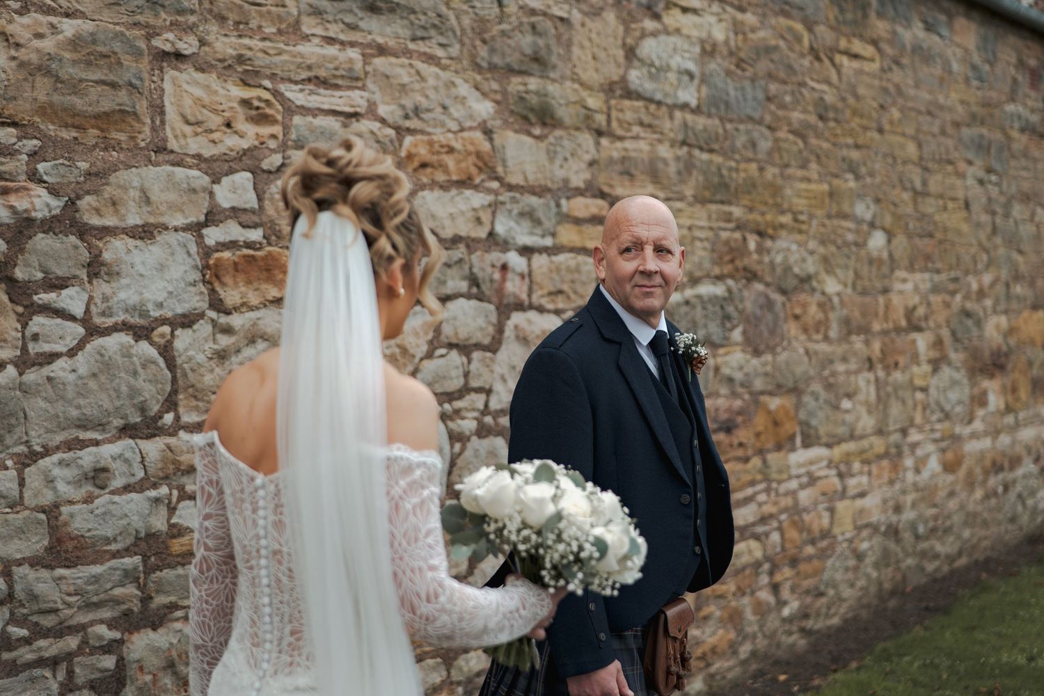 A couple stands against a rustic stone wall during a wedding ceremony with white floral arrangements.