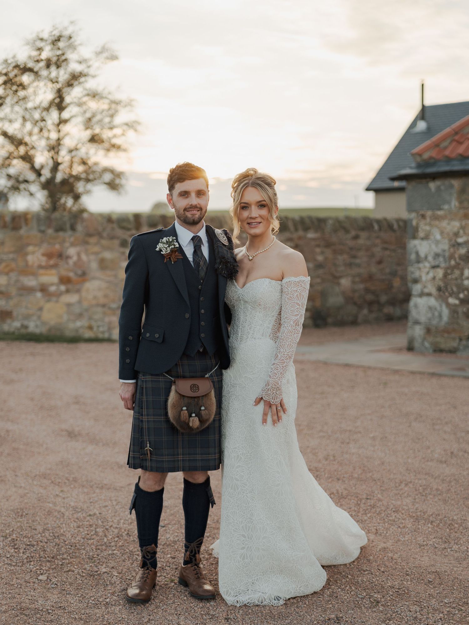 A couple in traditional Scottish wedding attire pose together on rustic grounds at sunset at Falside Mill.
