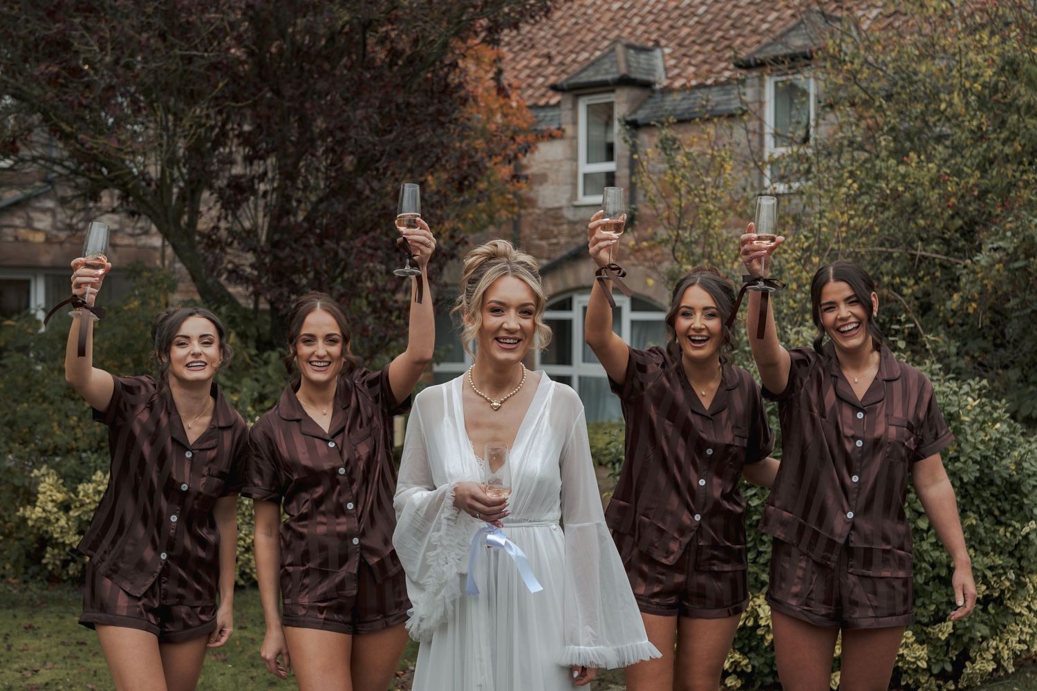 A group of friends in brown and white outfits pose and laugh together in an outdoor garden setting.