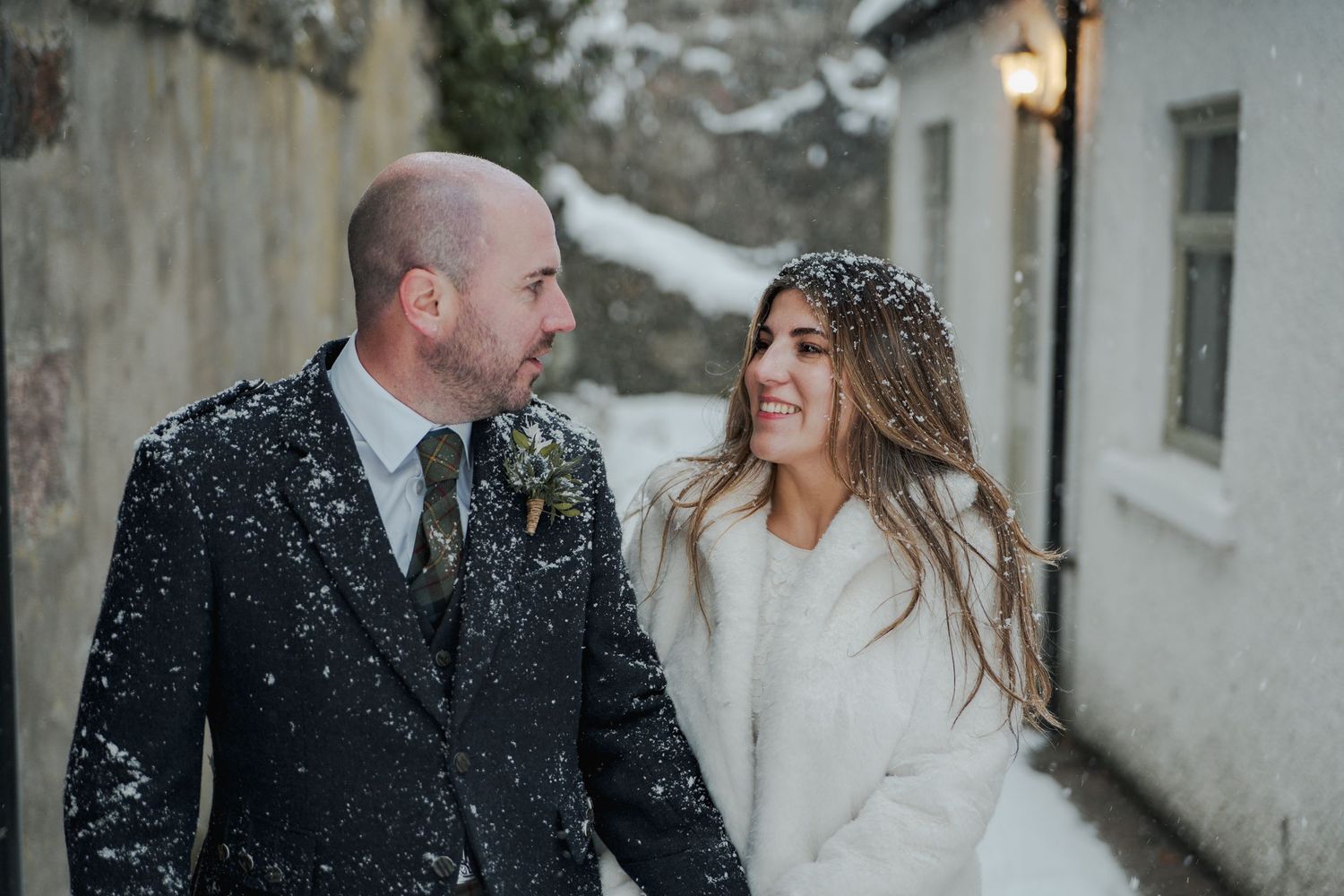 A happy couple shares a joyful moment in the snow, with the bride wearing a white winter coat over her wedding dress.