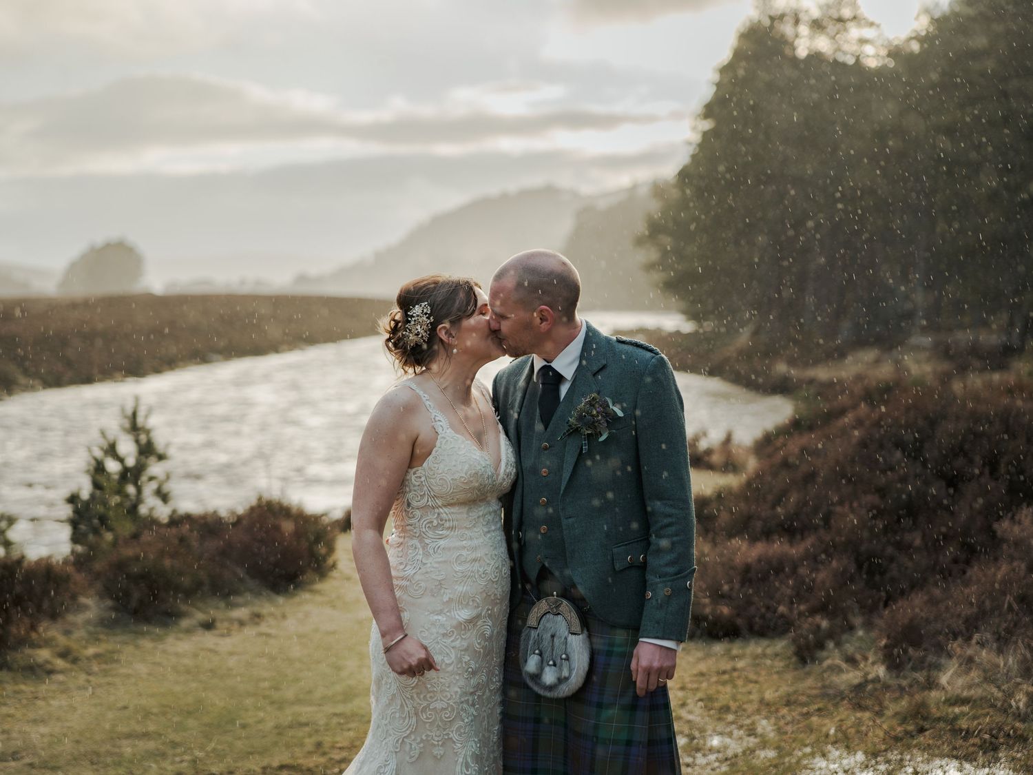 Couple sharing a romantic kiss in Scottish Highlands, groom in green tartan kilt, bride in lace dress, misty loch backdrop.