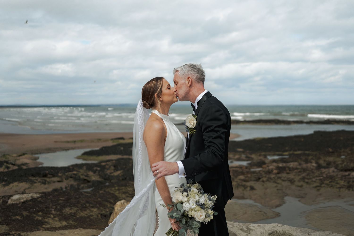 Bride and groom share a romantic kiss on rocky coastal cliffs with waves and cloudy skies behind them.