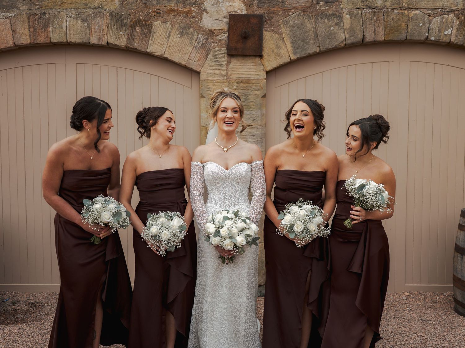 A wedding party poses in chocolate brown and white dresses holding white floral bouquets at Falside Mill.