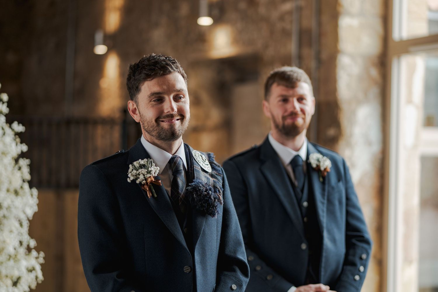 Groomsmen in black suits share emotional reactions during a rustic indoor wedding ceremony at Falside Mill.