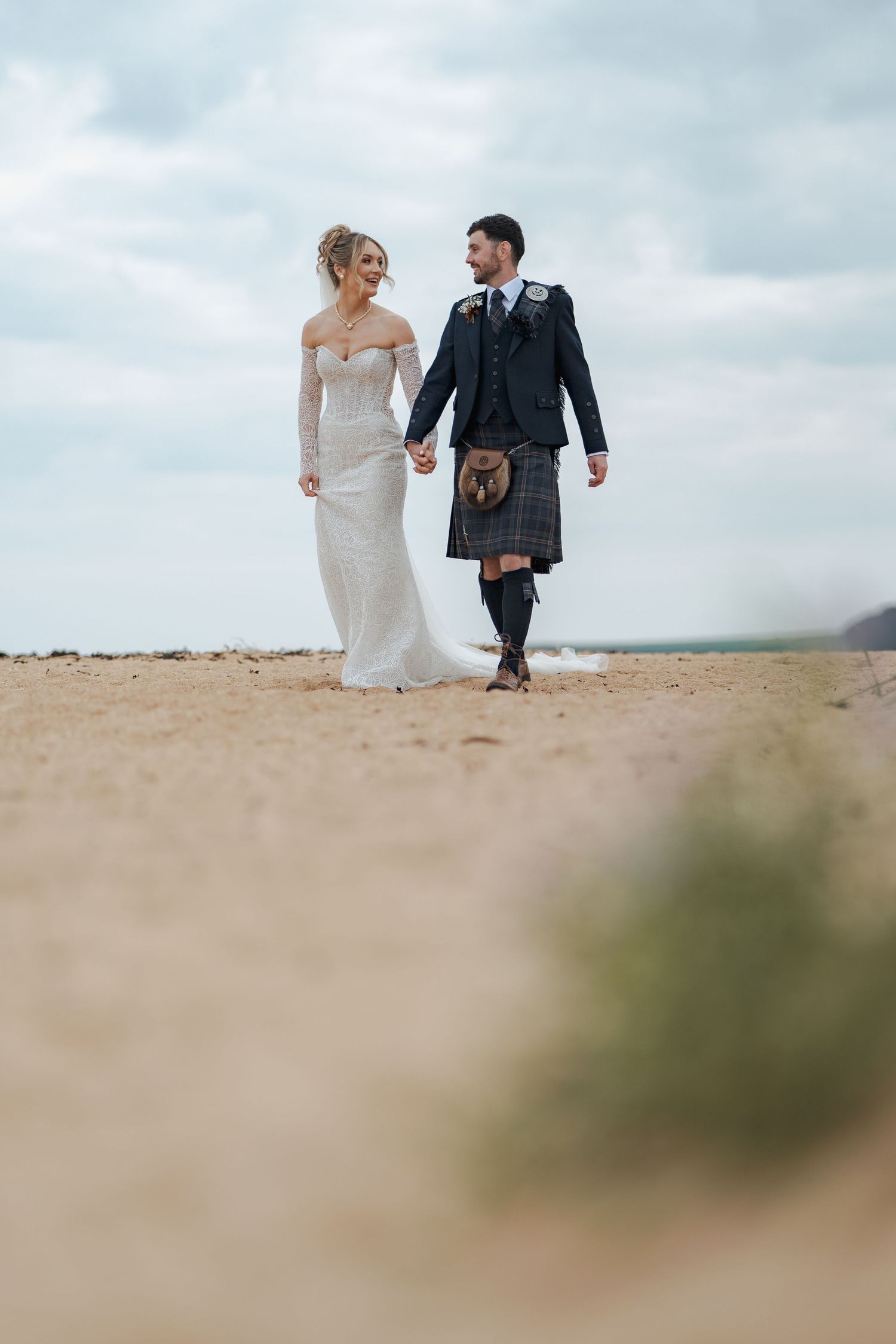 A bride in a white gown and groom in a suit walk together on a sandy Kingsbarns beach under cloudy skies.