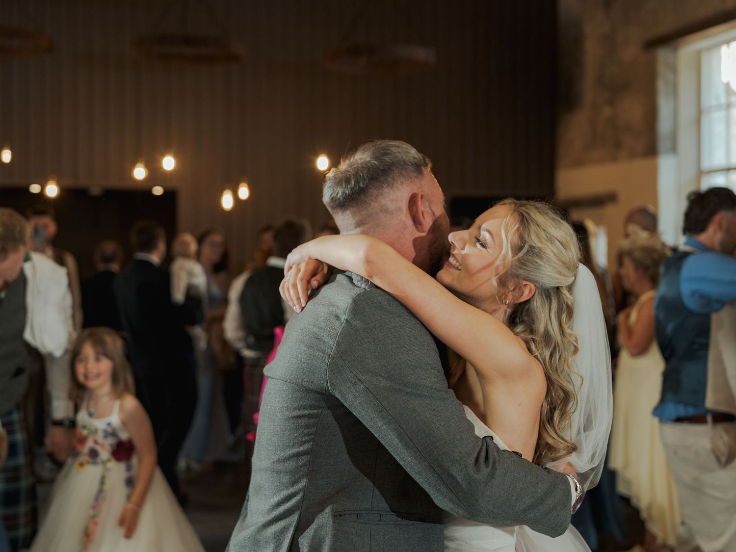Bride and groom share an emotional first dance embrace at rustic wedding reception with guests dancing in background.