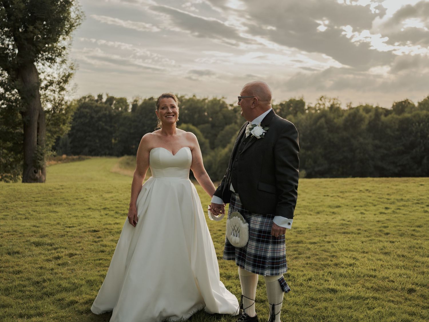 Bride in white gown and groom in blue tartan kilt hold hands on green lawn at outdoor wedding ceremony.