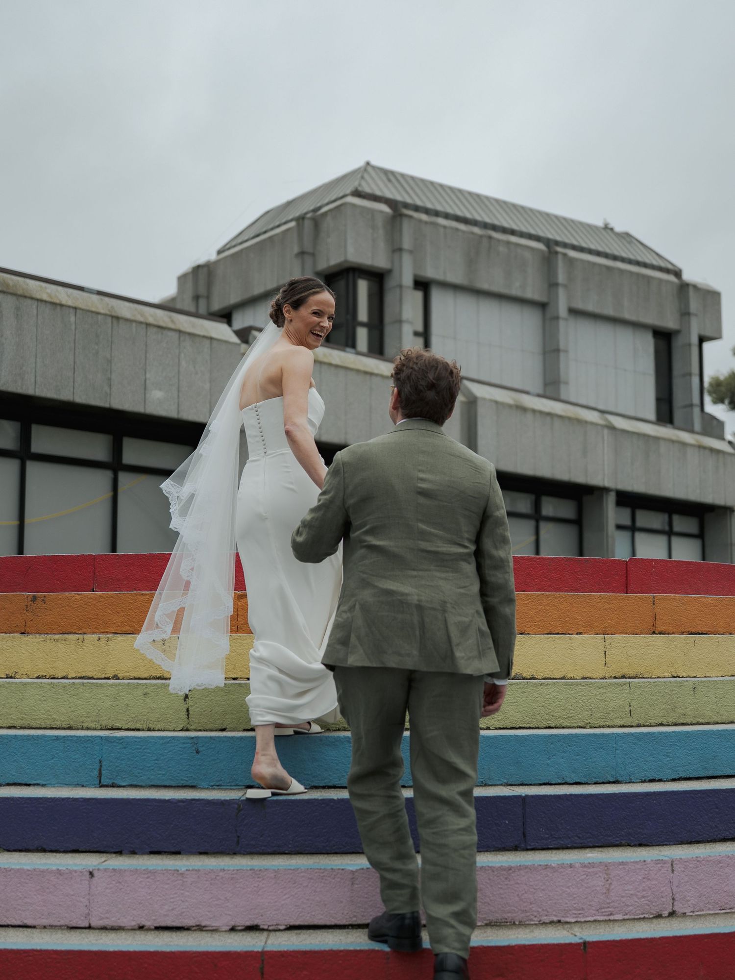 Bride in white gown and veil holds hands with groom in olive suit on rainbow-painted steps outside a brutalist building.