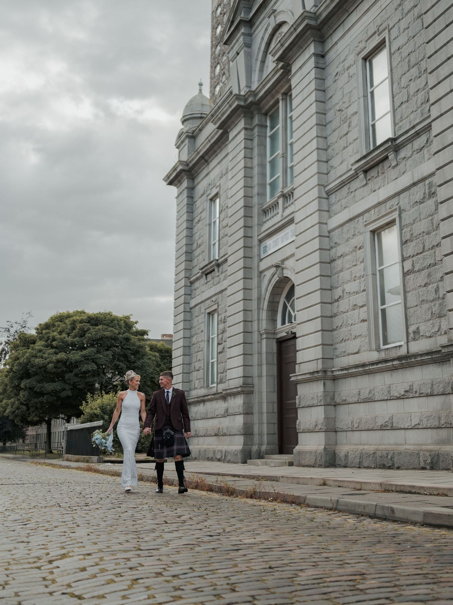 Bride and groom in Scottish kilt walking on cobblestone street beside grand granite church under cloudy sky.