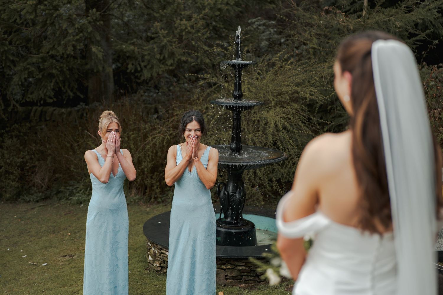 Two emotional bridesmaids in blue dresses react with joy seeing bride in white wedding gown near decorative fountain outdoors.