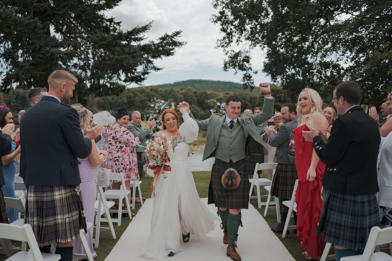 Joyful couple walks down outdoor wedding aisle with rolling hills in background while guests celebrate.