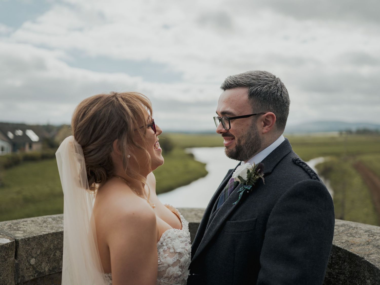 Bride and groom share a romantic moment on a stone wall with scenic Irish countryside and river in the background.