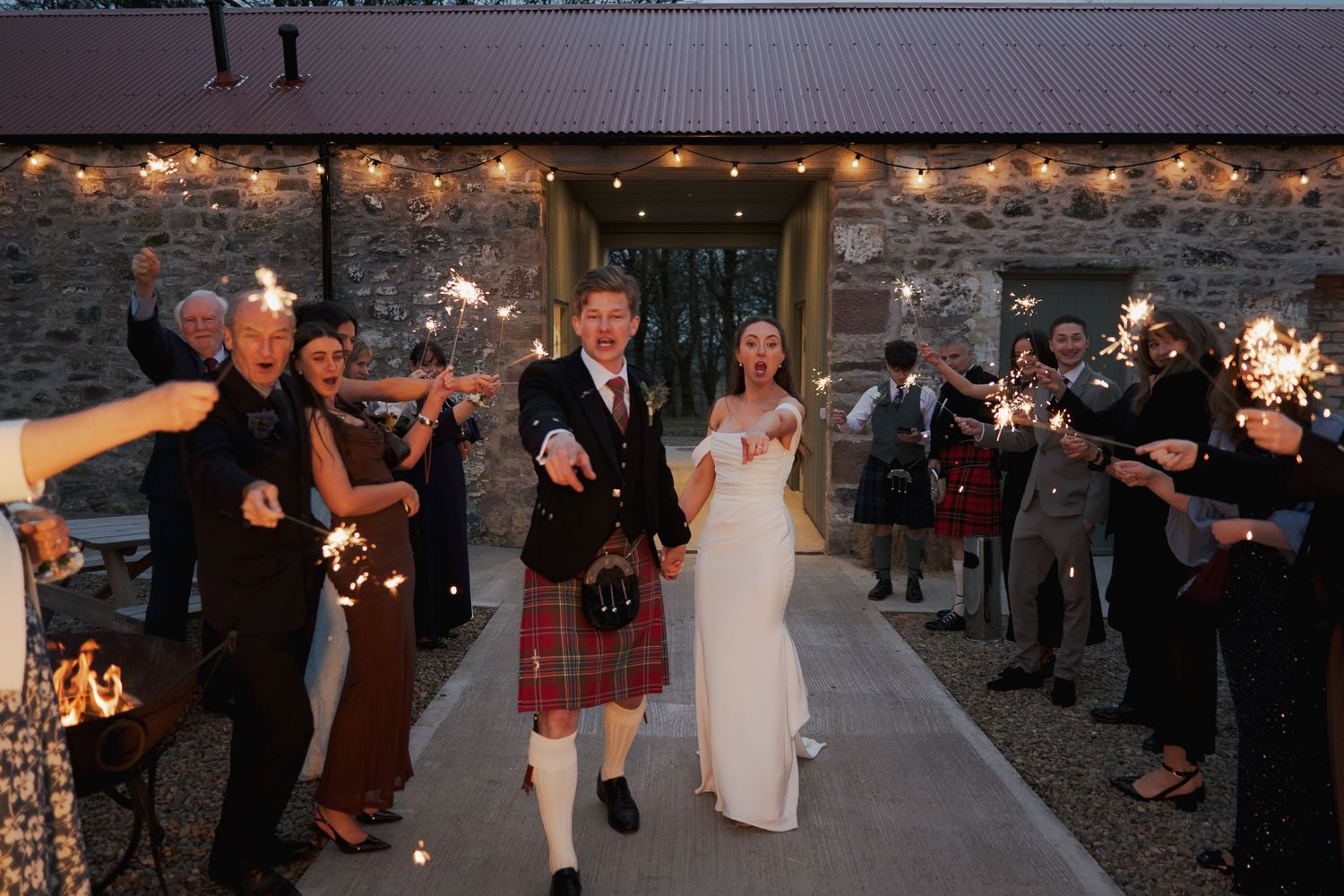 Bride and groom walk through sparkler send-off at Scottish wedding, groom wearing traditional tartan kilt.