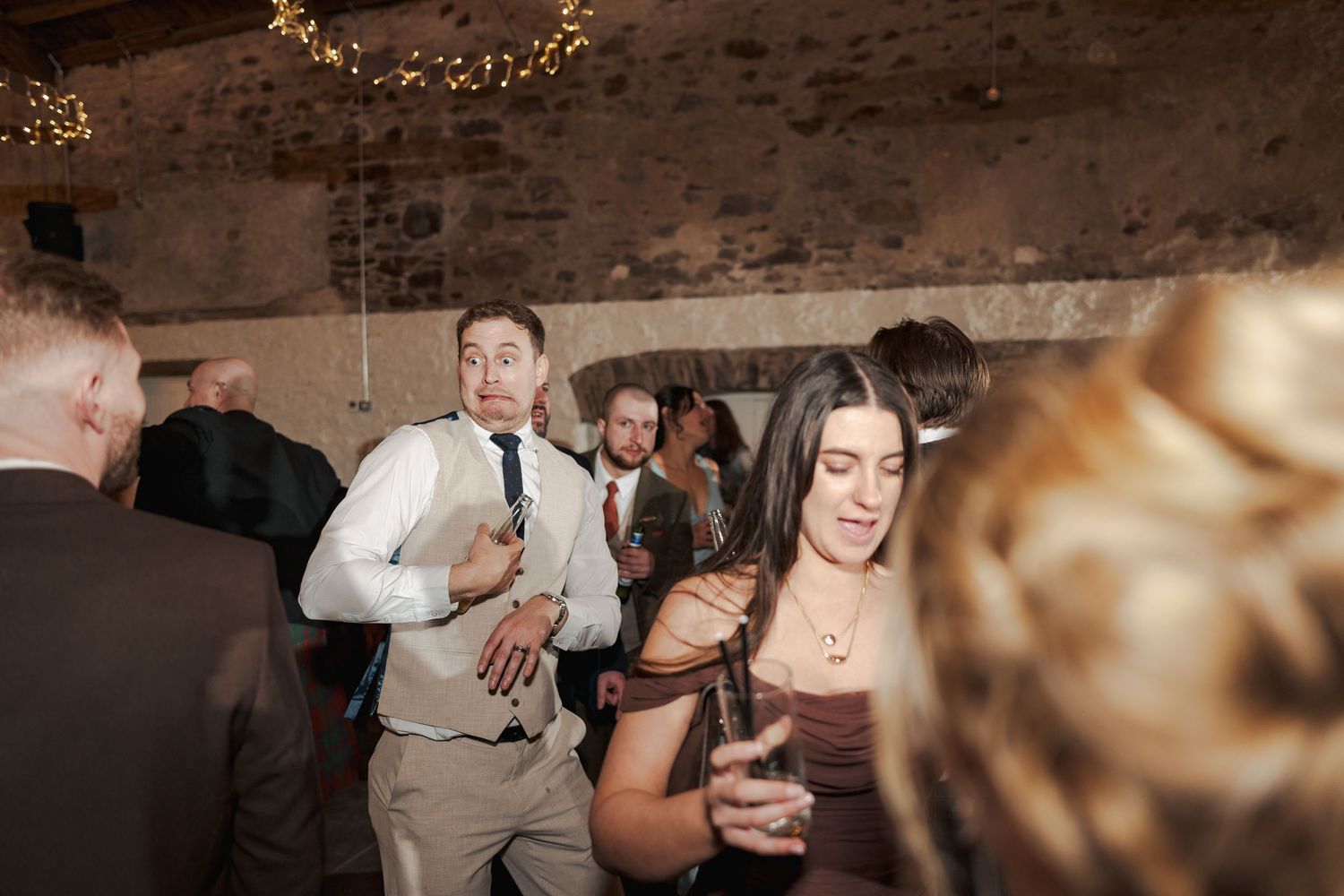 Guests dancing and socializing at a wedding reception in a rustic venue with string lights.