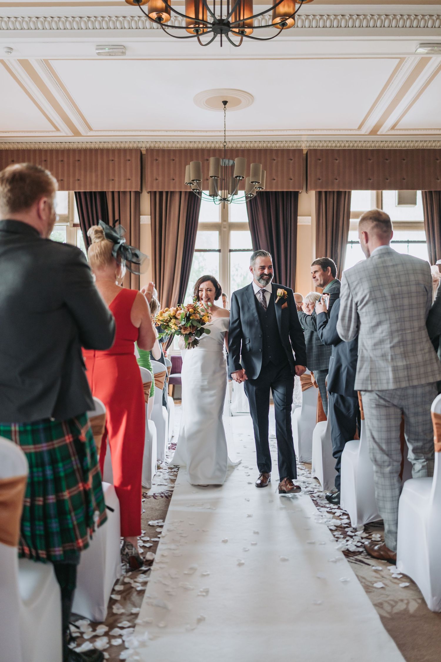 Wedding ceremony indoors at Dunblane Hydro with couple walking down white aisle runner while photographer captures moment in elegant venue.