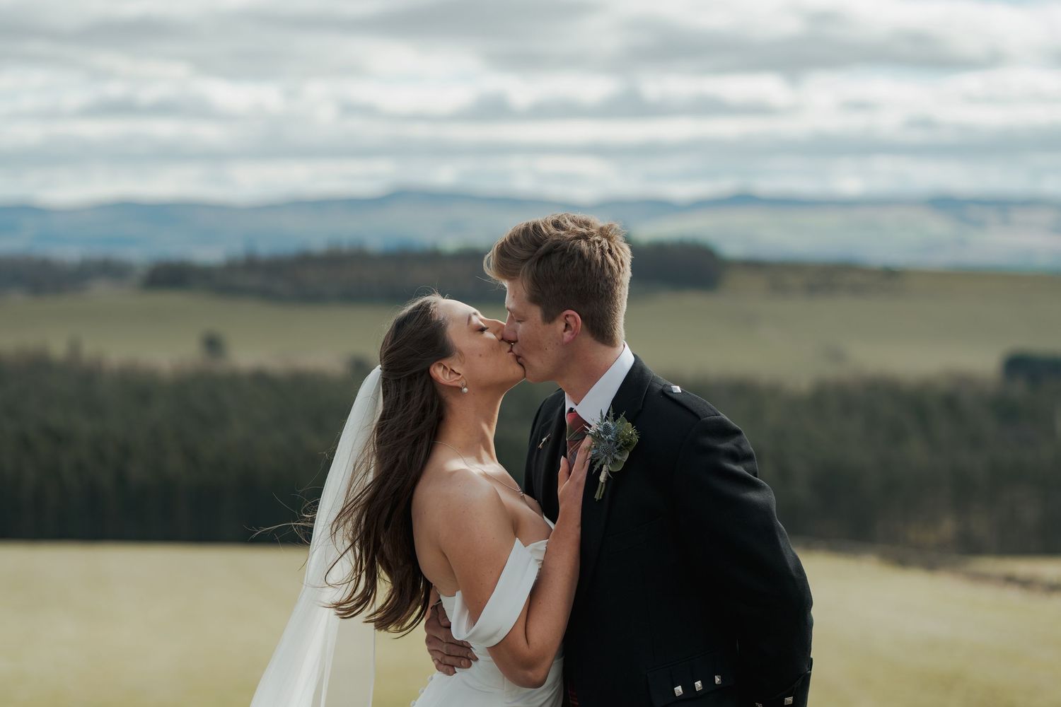 A bride and groom share a romantic kiss outdoors with scenic green fields and rolling hills in the background.
