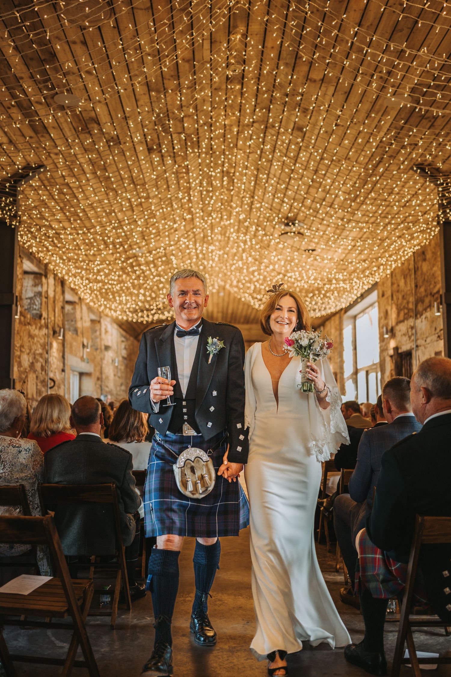 Newlyweds walking down aisle under stunning ceiling of fairy lights in Falside Mill.