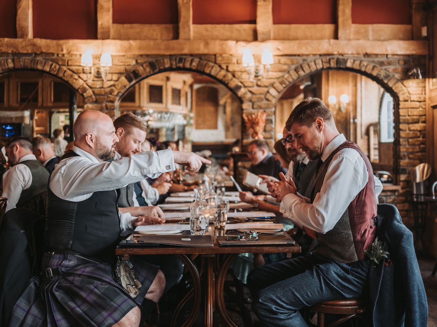 Wedding guests in traditional Scottish attire dining at a long table in rustic venue.