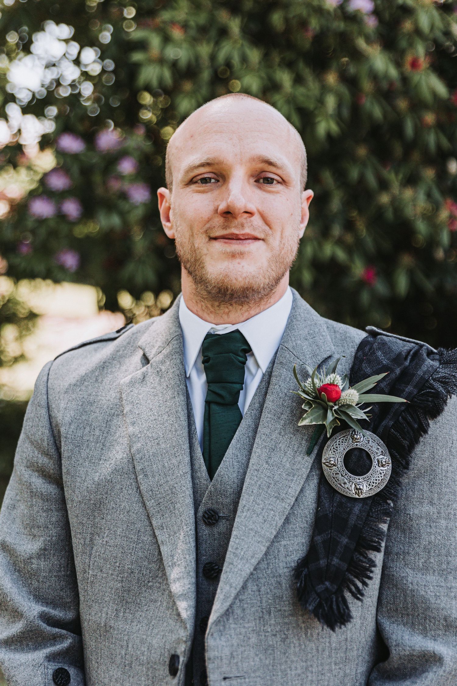 Groom in gray three-piece suit with dark tie and red boutonniere poses outdoors against garden background at Murrayshall.