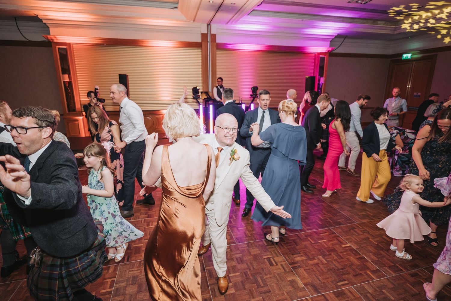 Wedding guests dance and celebrate on a brick-patterned floor under warm orange and purple venue lighting.