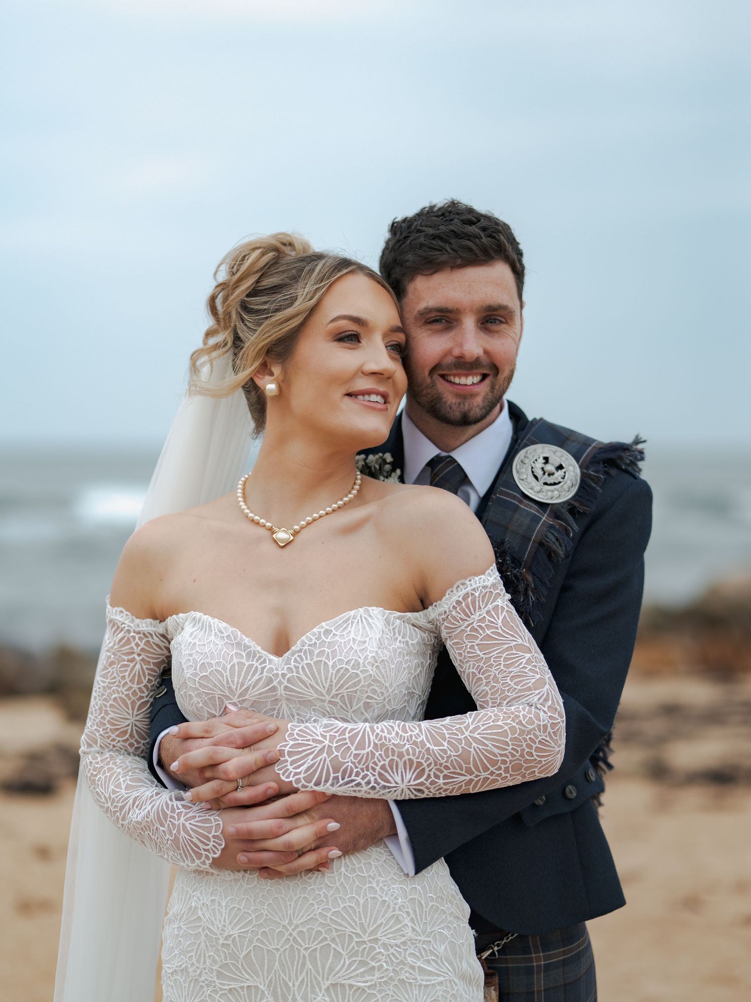 A couple shares intimate moments on Kingsbarns beach during their wedding photoshoot, showcasing a long-sleeved lace bridal gown.