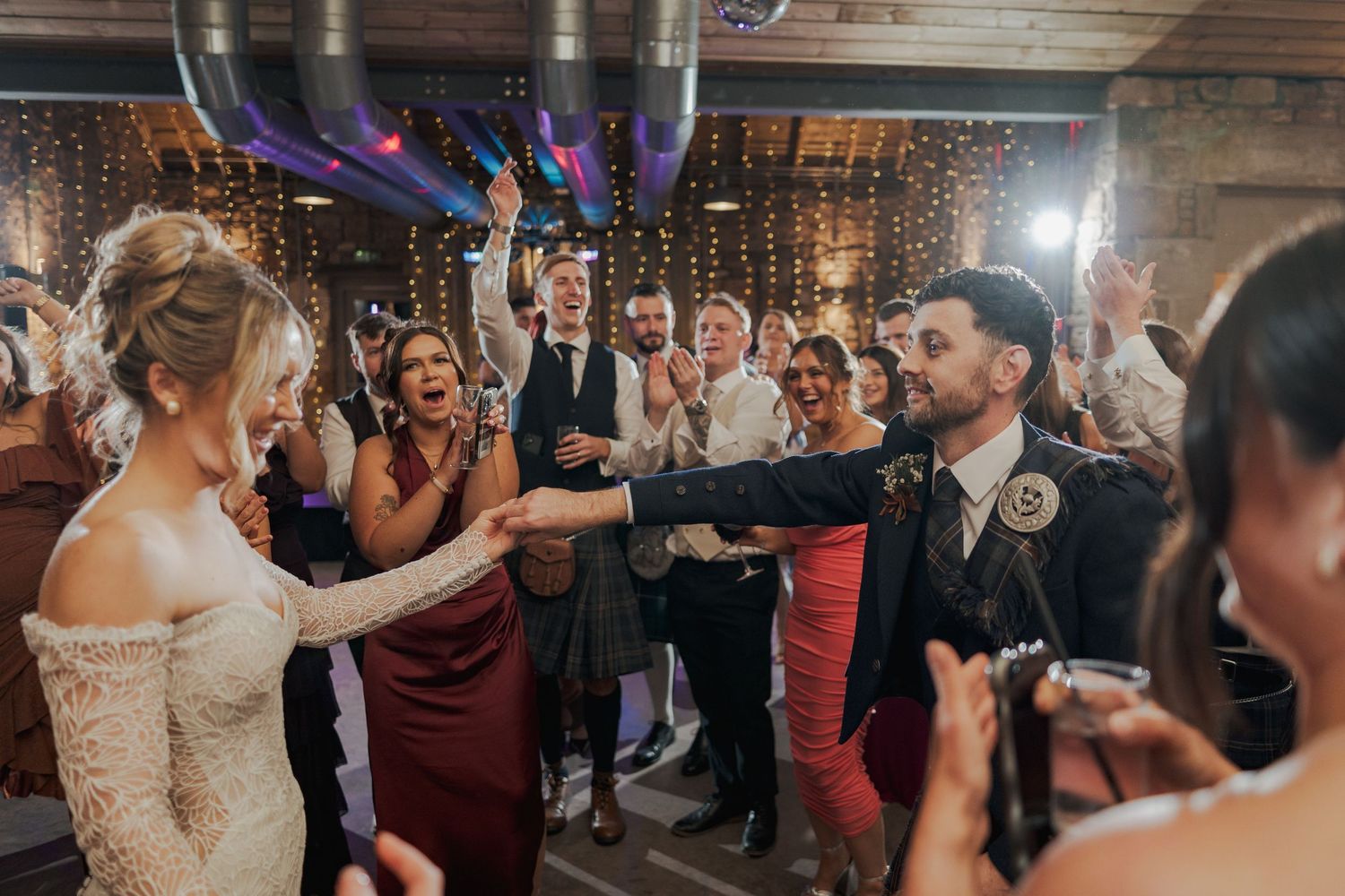 Wedding guests dance and celebrate together at Falside Mill on a crowded dance floor at a reception venue with purple lighting.