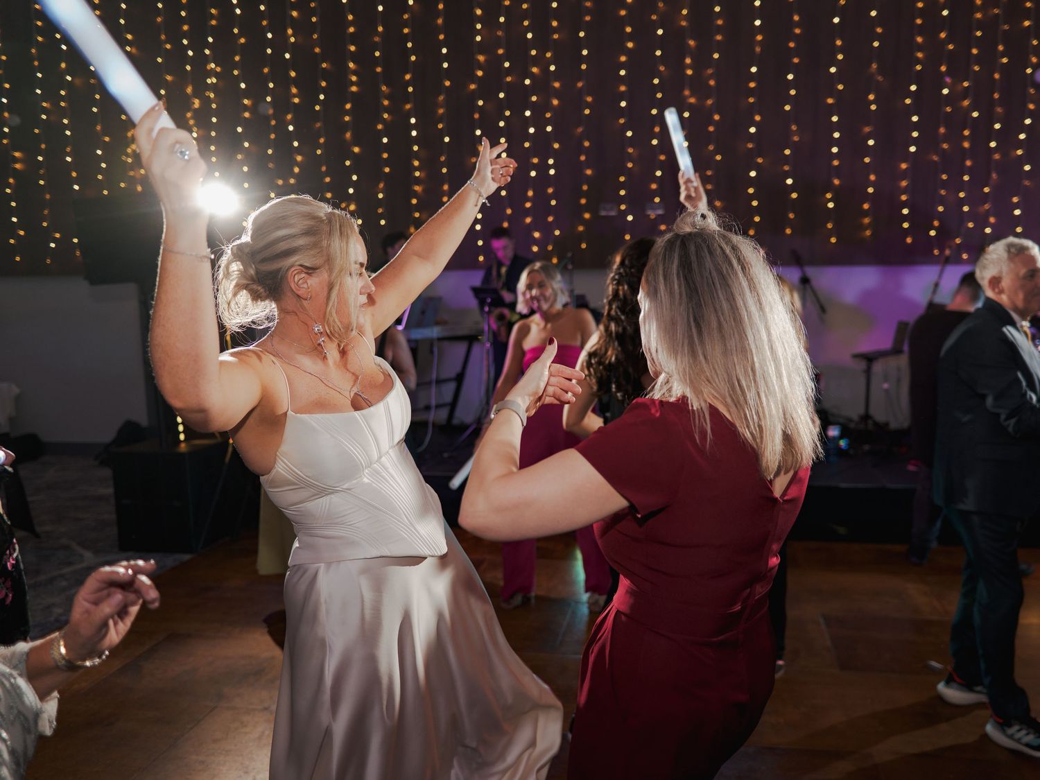 Wedding party dancing on a wooden dance floor beneath twinkling string lights during evening celebration.