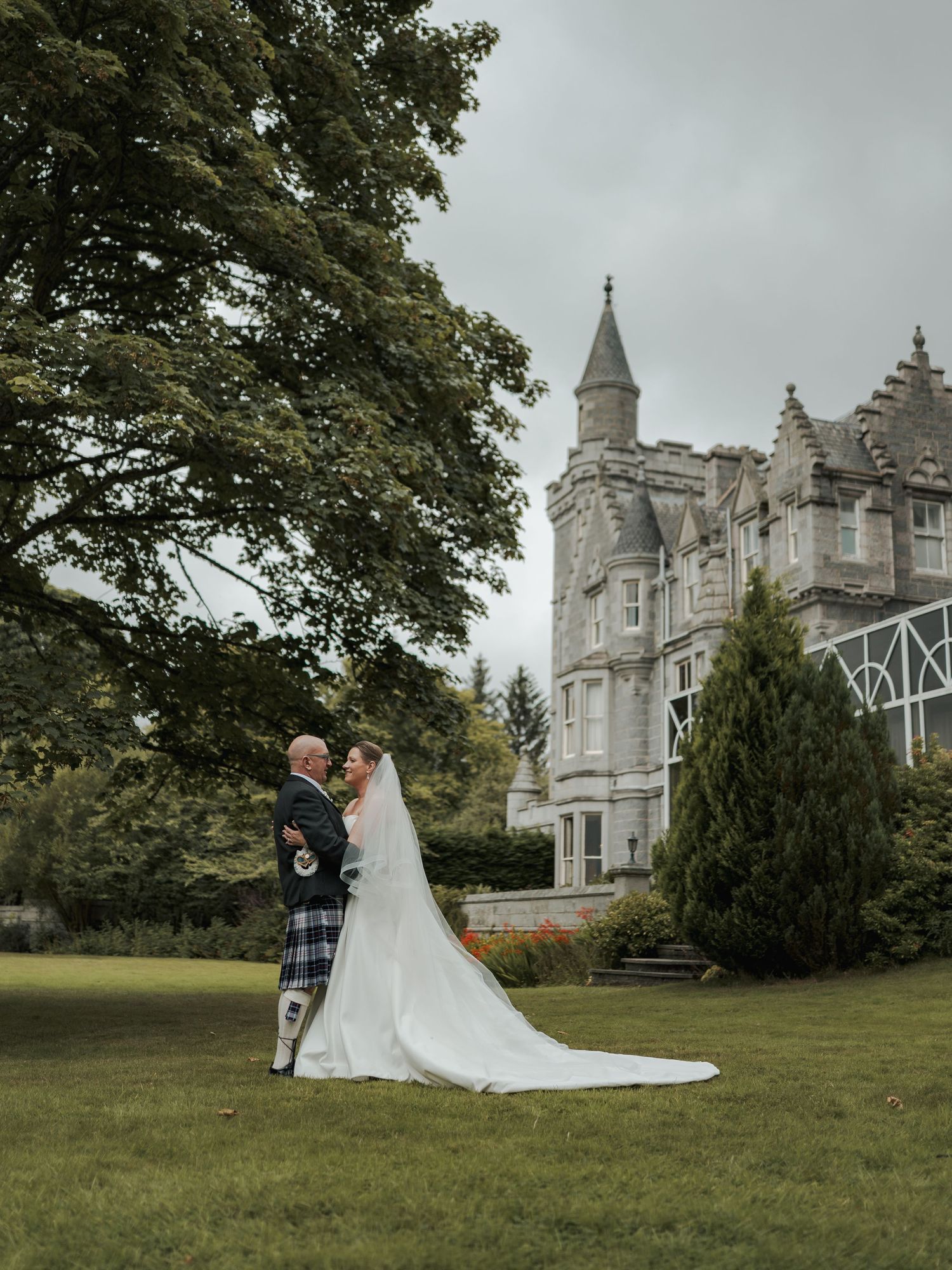A bride and groom share a romantic moment on the lawn of a Scottish castle, with the groom wearing a traditional tartan kilt.
