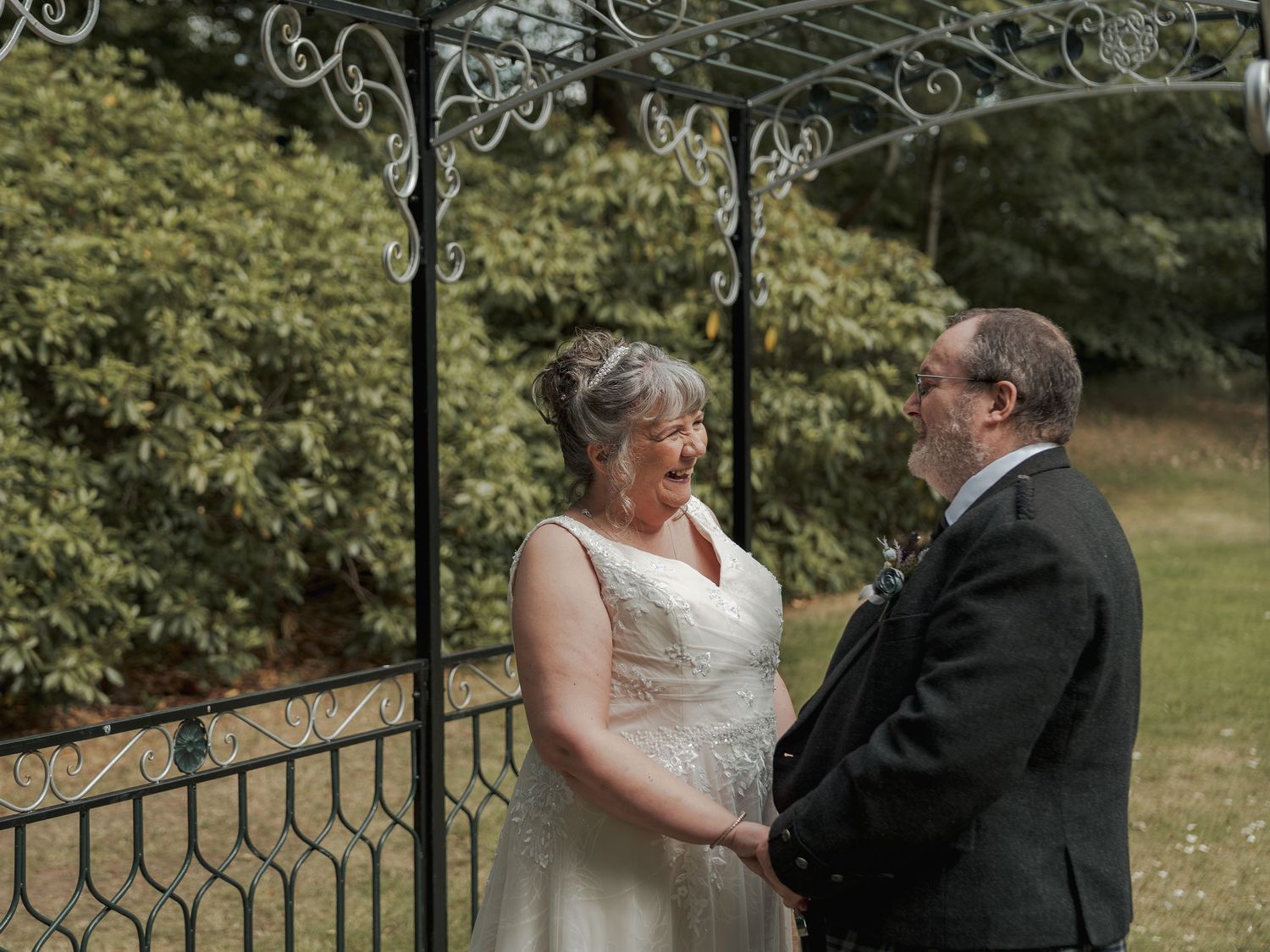Couple exchanging vows outdoors under decorative iron arbor, bride in white gown laughing joyfully with groom in dark suit.