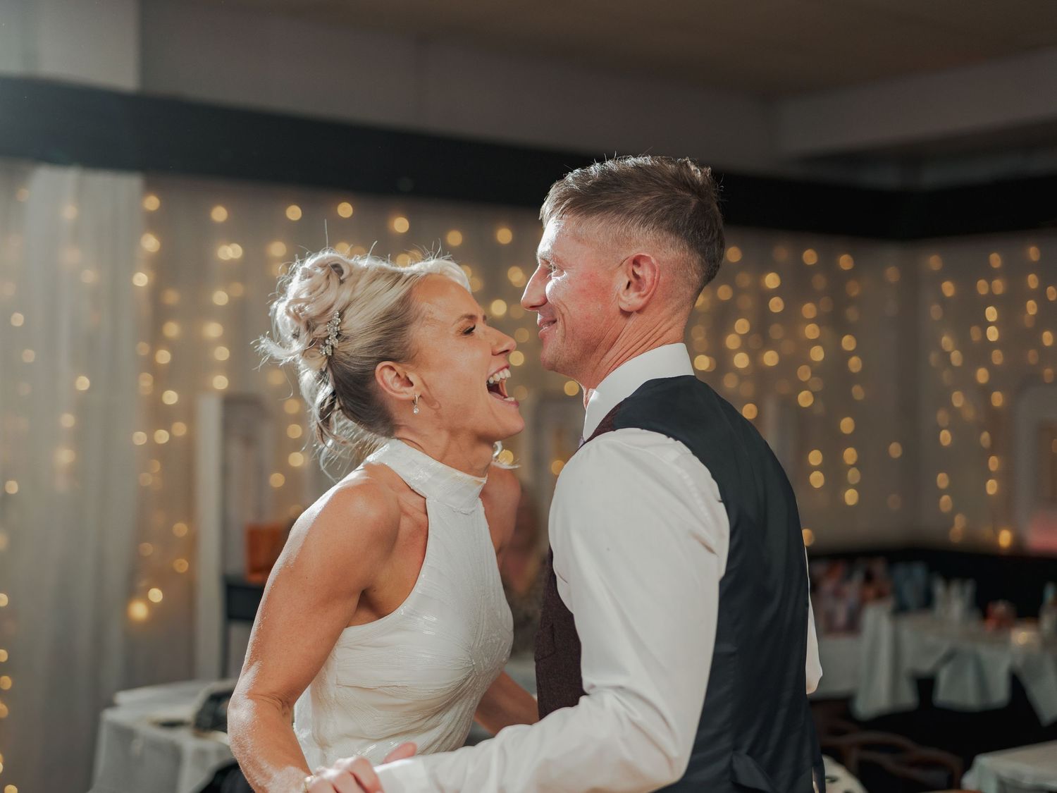 A joyful couple shares their first dance at a wedding reception, surrounded by warm fairy lights in the background.