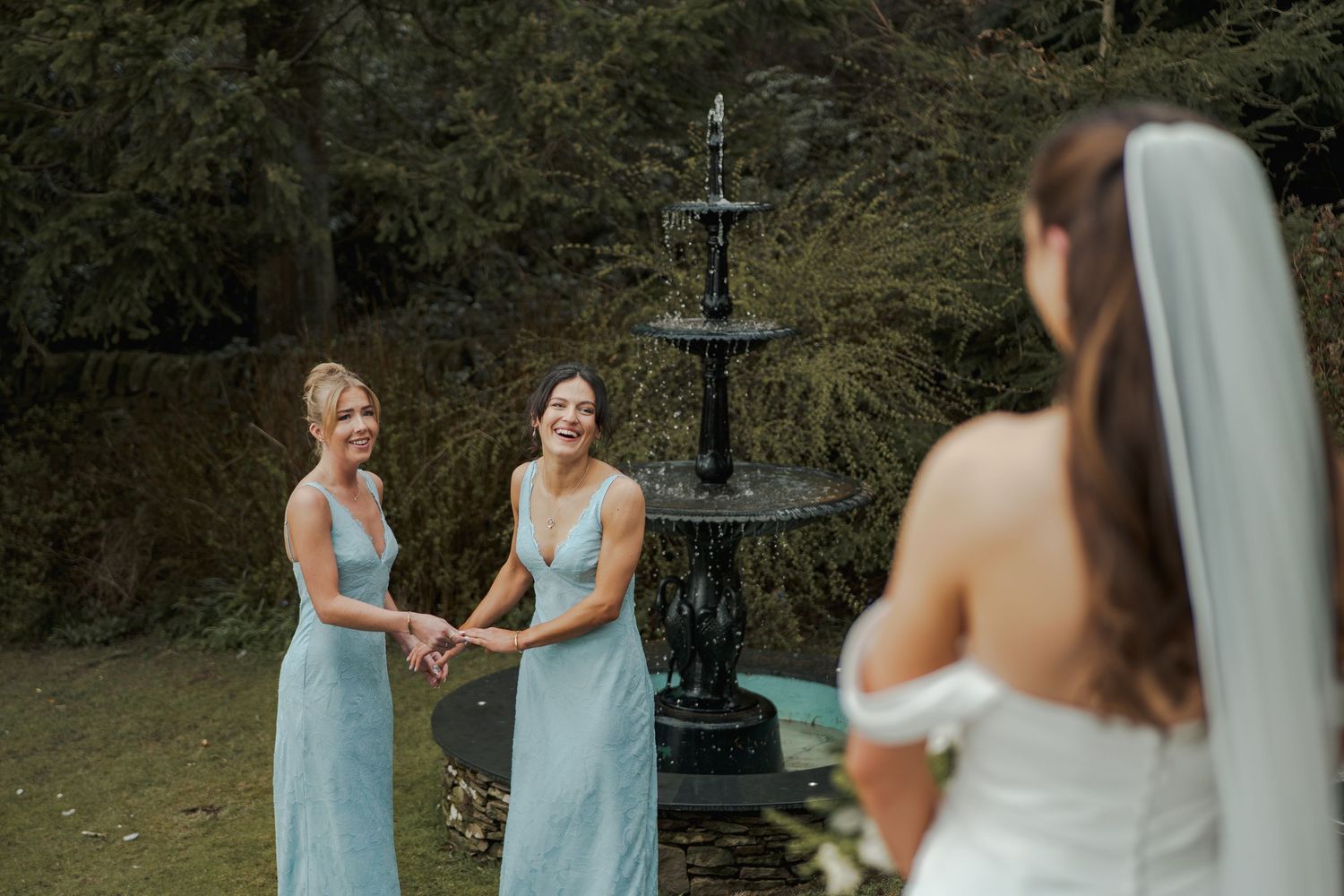 Bride in white gown and veil faces two smiling bridesmaids in light blue dresses near a black fountain outdoors.
