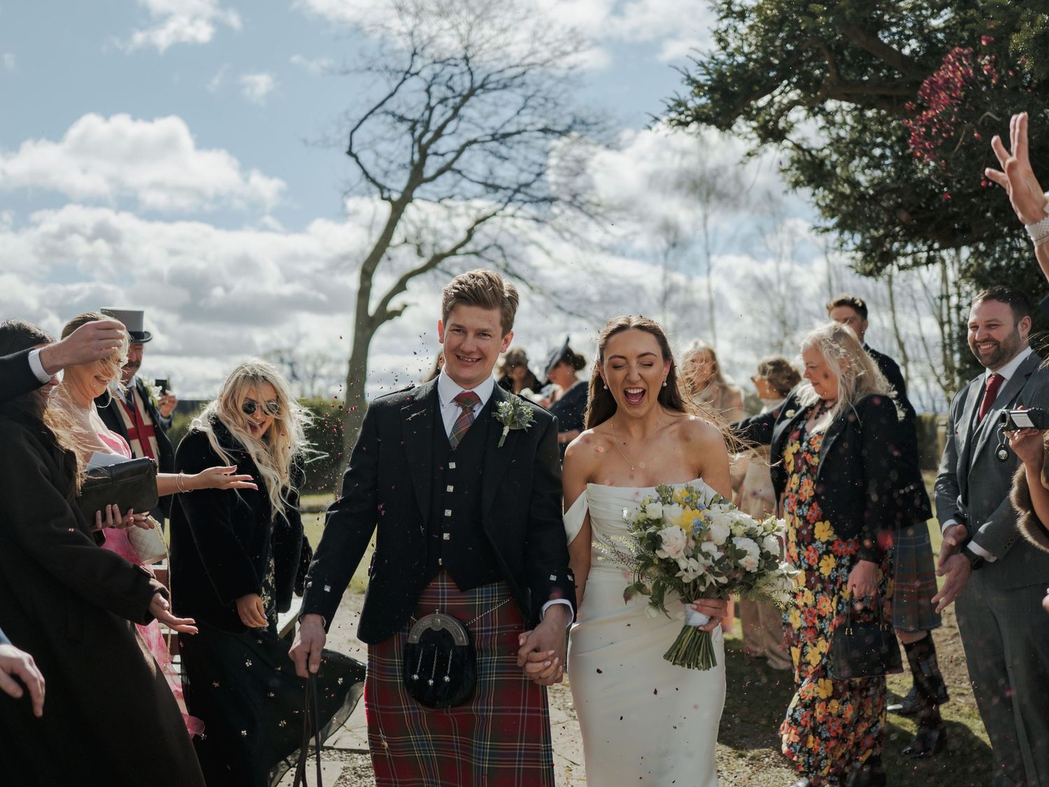 Newlyweds walk through confetti shower, groom in Scottish tartan kilt, bride in white gown holding bouquet, guests cheering outdoors.