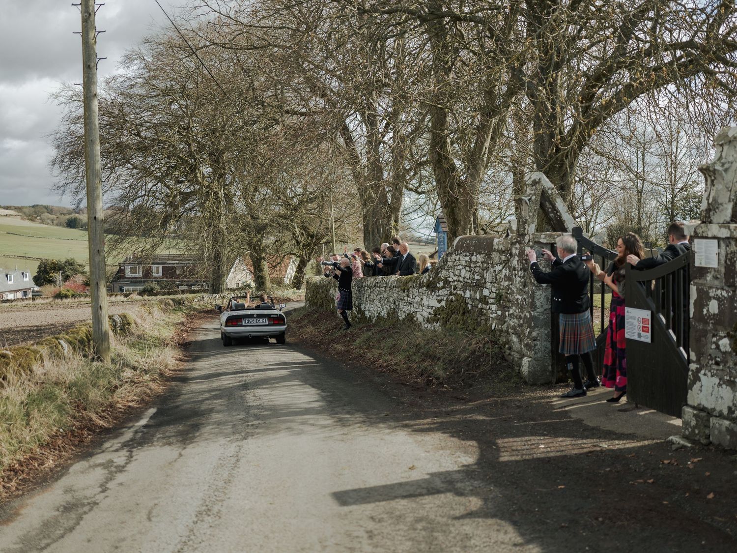 Spectators line a rural stone wall road watching a car pass during a countryside rally event on a cloudy day.