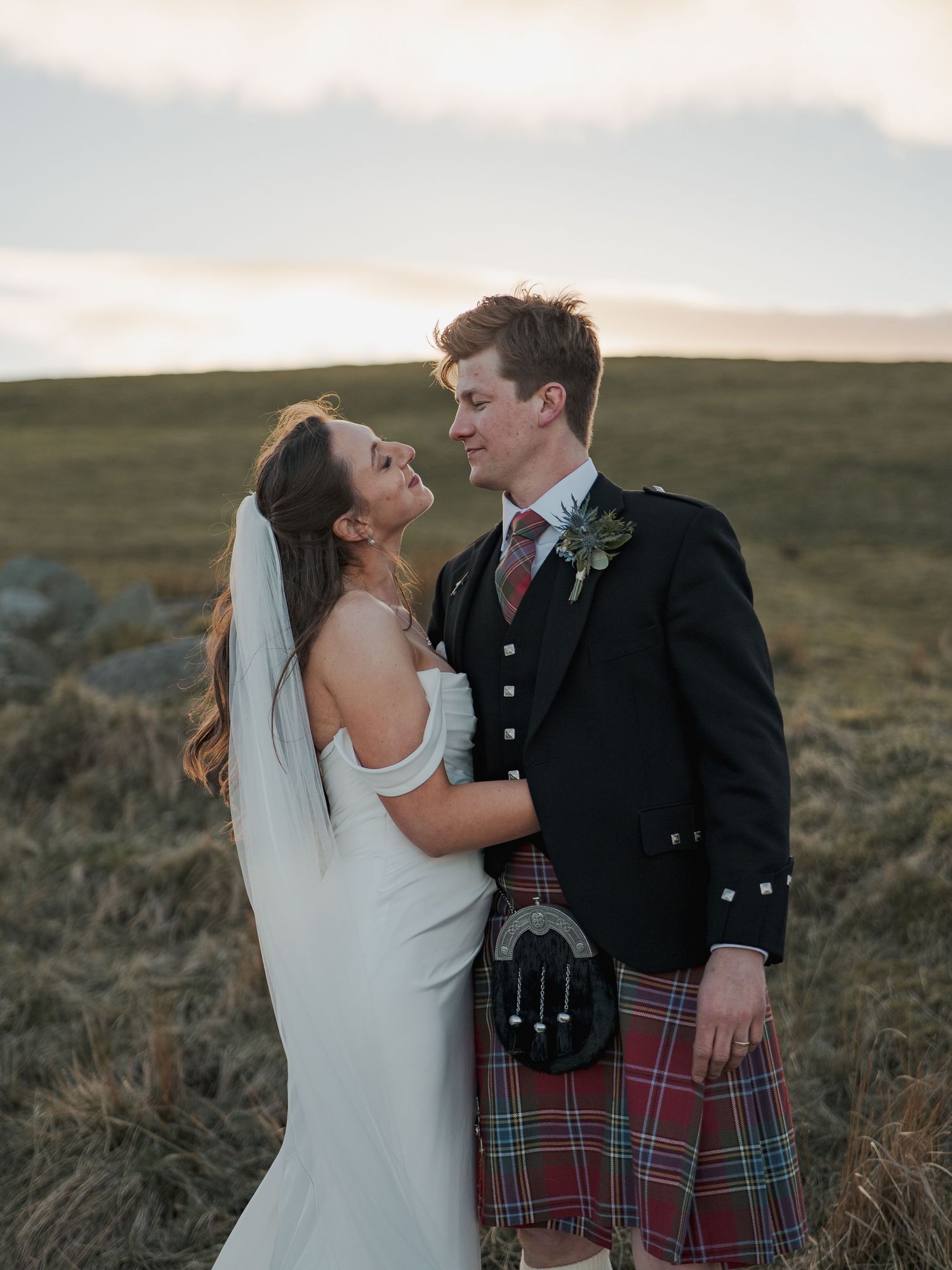 A bride in a white gown and groom in a Scottish kilt share a romantic moment in an open highland landscape.