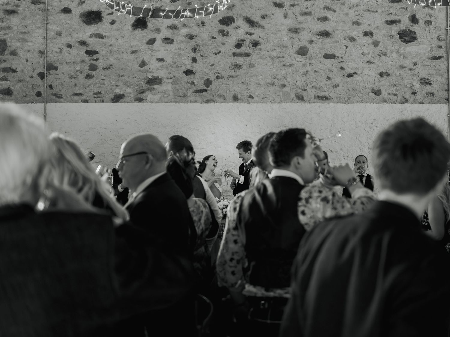 Black and white photo of elegantly dressed wedding guests mingling in a rustic stone-walled venue.