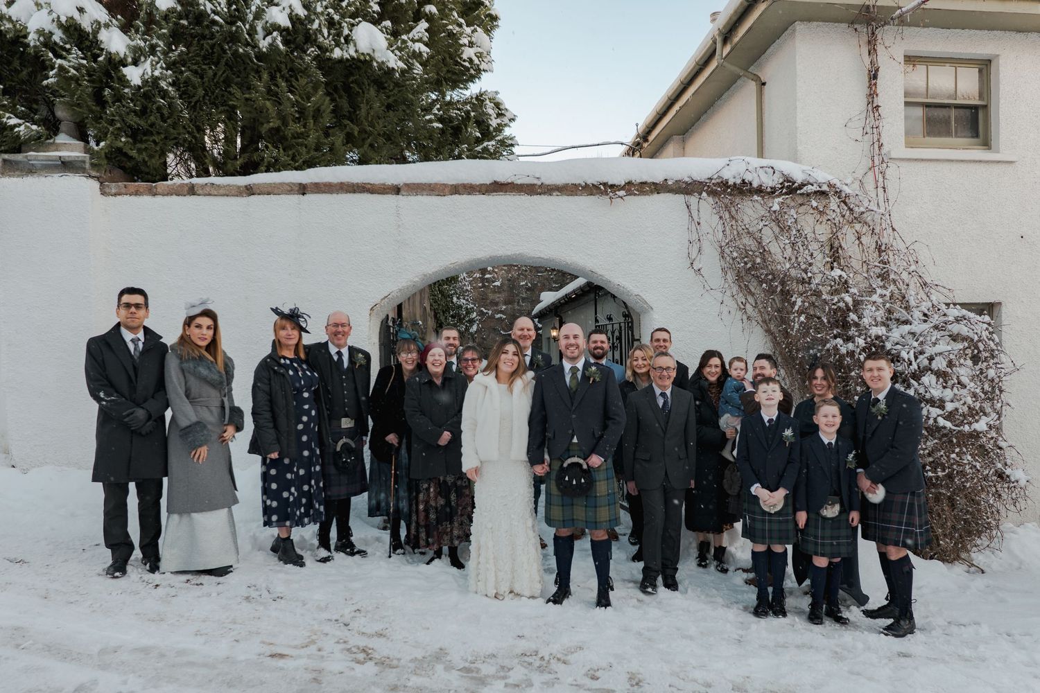 Wedding group photo in snowy winter setting, guests in formal attire stand before stone archway.