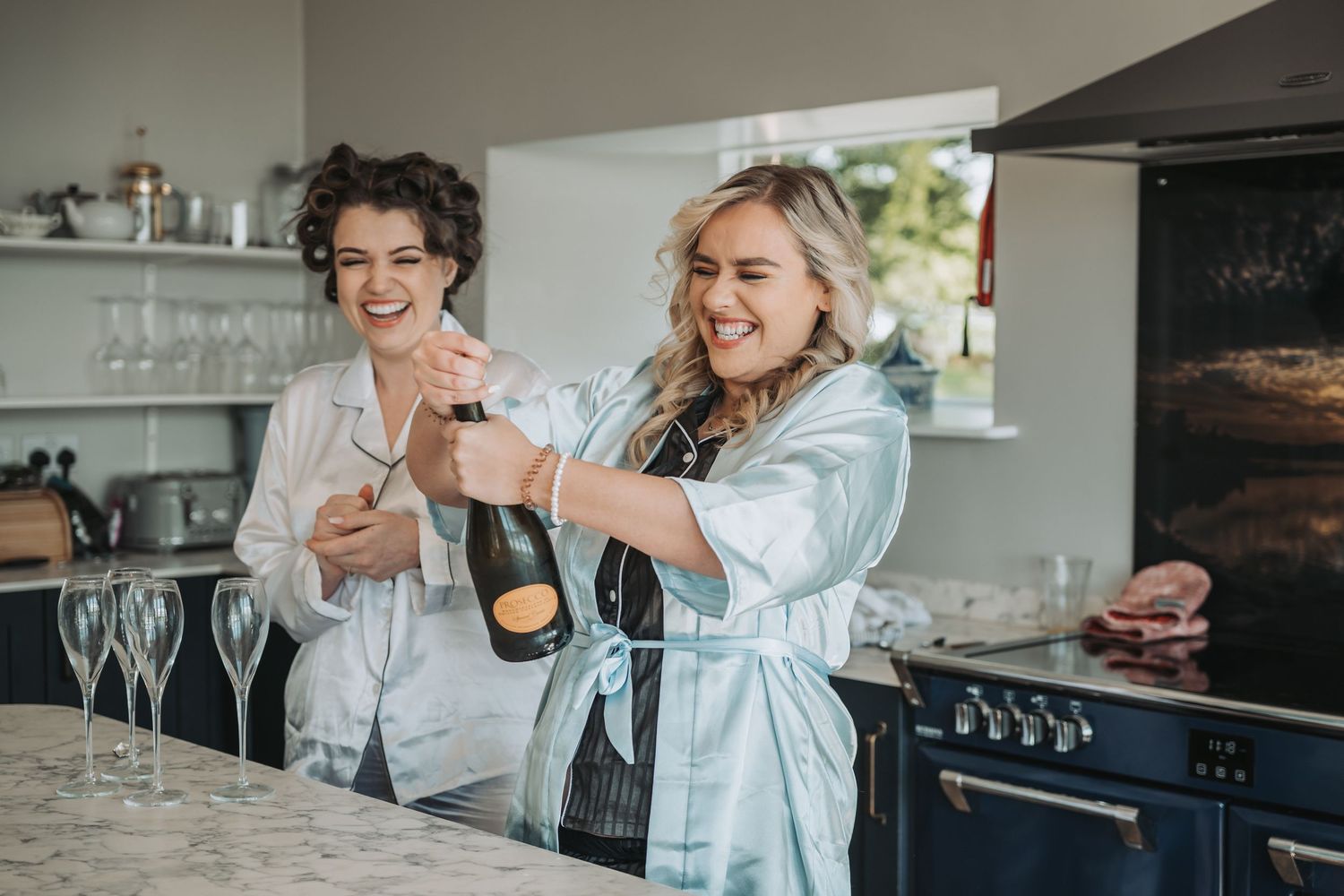 Two people in silk robes open a champagne bottle at a marble kitchen counter with champagne flutes nearby.