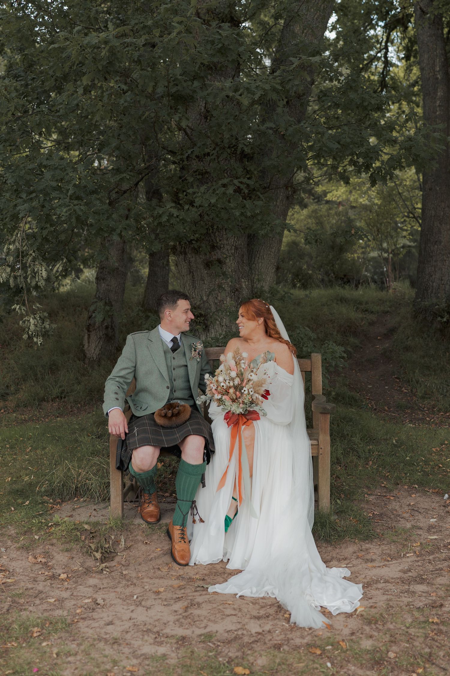 A couple sits together on a rustic wooden bench surrounded by a wooded forest area during an autumn wedding.